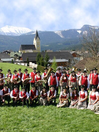 A large group of people in traditional attire poses on a meadow. In the background, mountains and a village can be seen. | © Musikkapelle Bad Mitterndorf