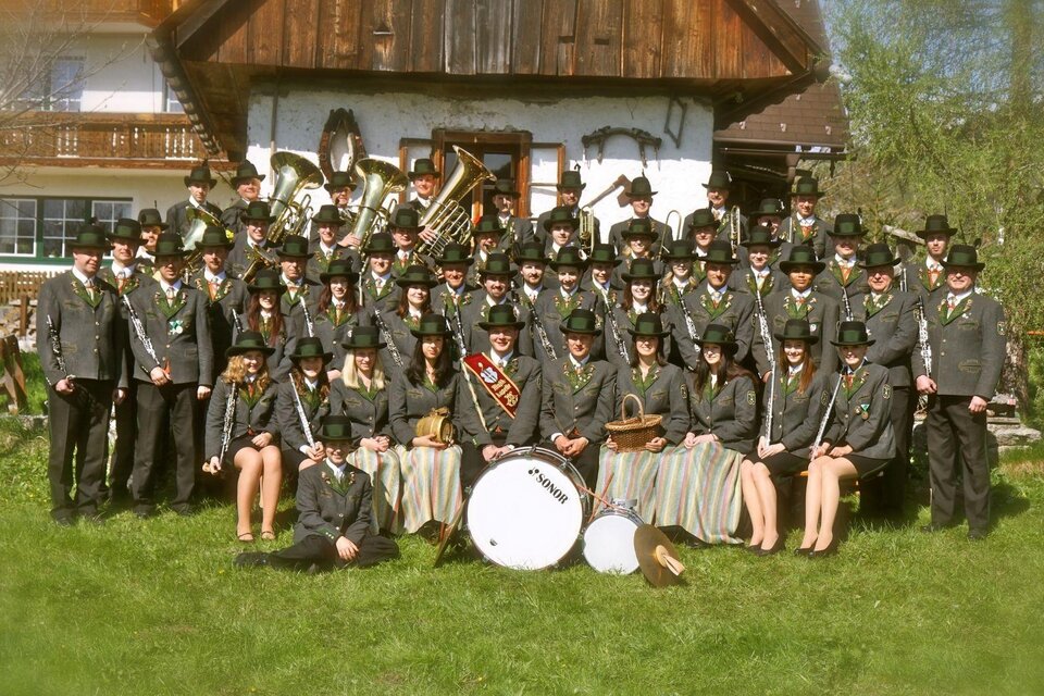 Eine große Gruppe von Menschen in traditionellen Trachten steht vor einem rustikalen Haus. Viele tragen Hüte und halten Musikinstrumente. | © Musikkapelle Kumitz