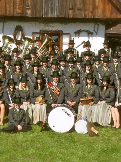 A large group of people stands in front of a traditional house. They wear festive clothing and hats, while some musical instruments can be seen. | © Musikkapelle Kumitz