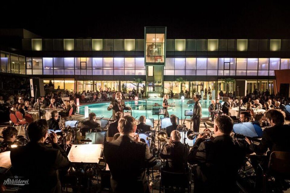 Die Big Band Köflach spielt in der Therme NOVA, im Hintergrund ein Thermalbecken und das Thermengebäude. | © Die Abbilderei