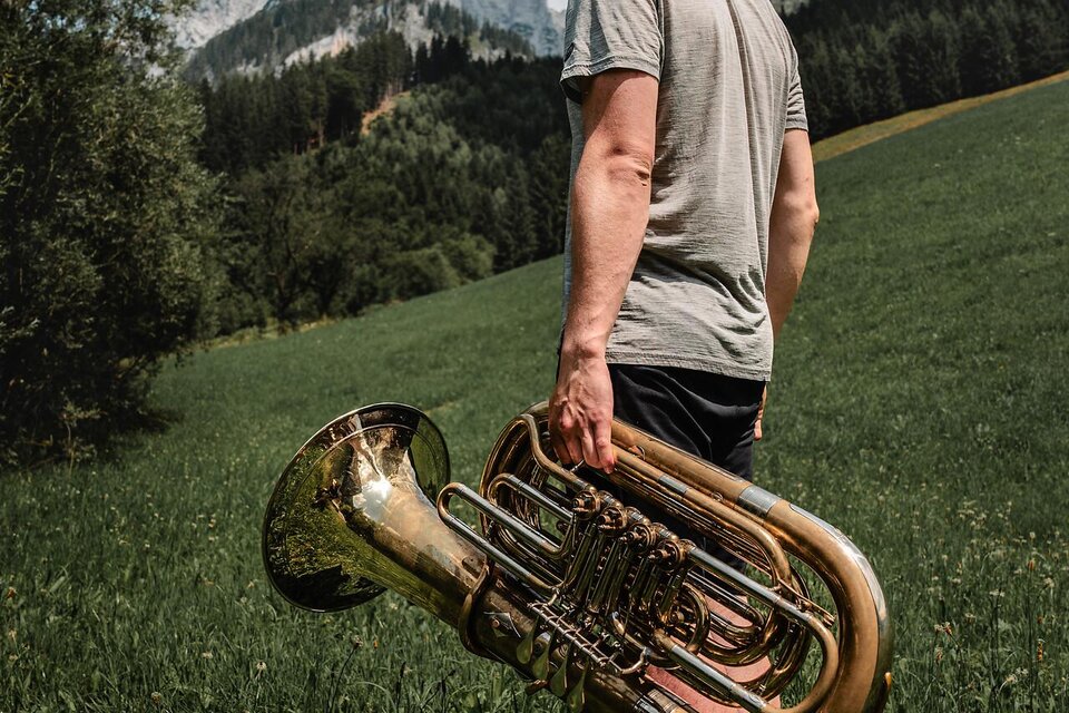 Ein junger Mann steht in einer grünen Wiese und hält eine Tuba. Im Hintergrund sind Berge und ein blauer Himmel zu sehen. | © TV Gesäuse