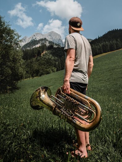 Ein junger Mann steht in einer grünen Wiese und hält eine Tuba. Im Hintergrund sind Berge und ein blauer Himmel zu sehen. | © TV Gesäuse