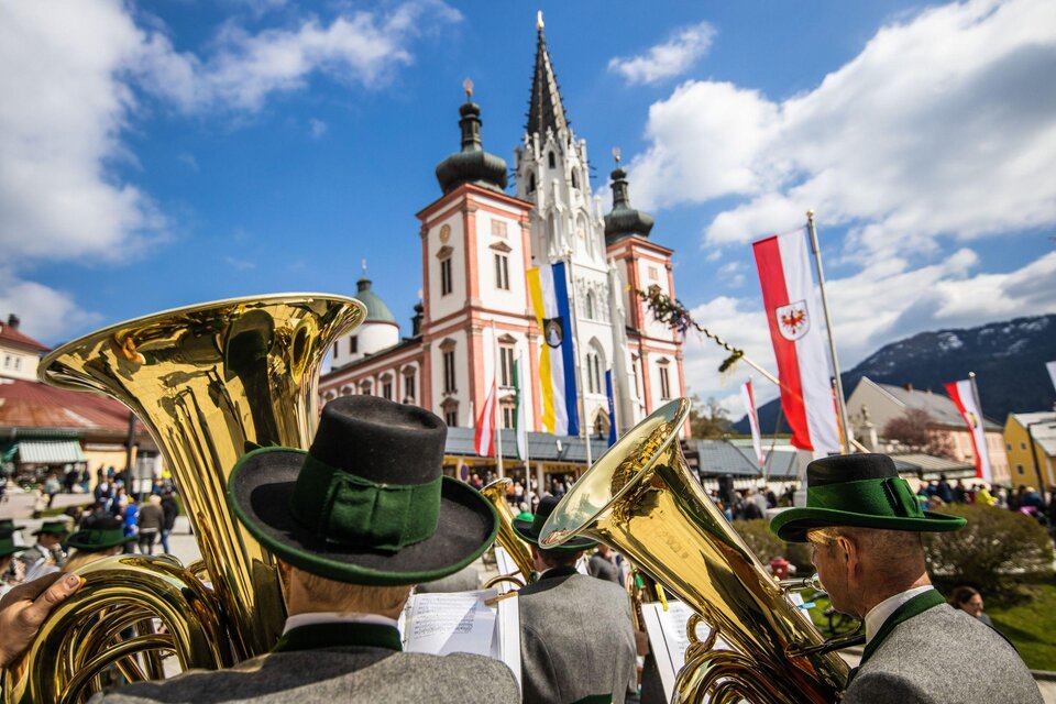 Eine Blaskapelle spielt vor einem historischen Gebäude mit Fahnen und Bergen im Hintergrund. Es ist ein sonniger Tag mit einigen Wolken. | © Fred Lindmoser