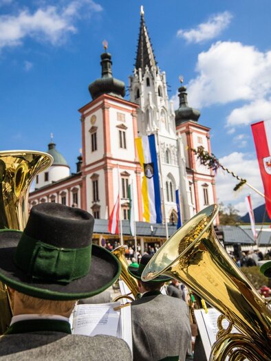Eine Blaskapelle spielt vor einem historischen Gebäude mit Fahnen und Bergen im Hintergrund. Es ist ein sonniger Tag mit einigen Wolken. | © Fred Lindmoser