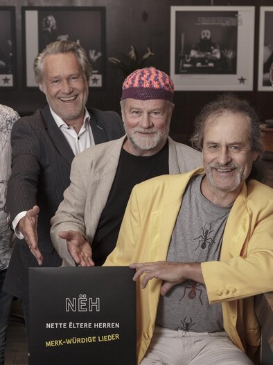 Four men are standing at a table and smiling at the camera. They are presenting an album titled "NÉH – Nice Older Men." | © Weinberger