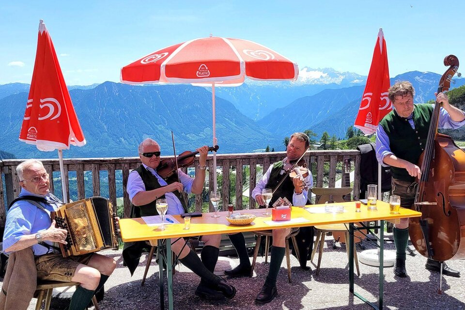 Eine Gruppe von Musikern spielt traditionelle Instrumente unter einem Sonnenschirm mit Blick auf die Berge. Die Stimmung ist fröhlich und einladend. | © RHuber