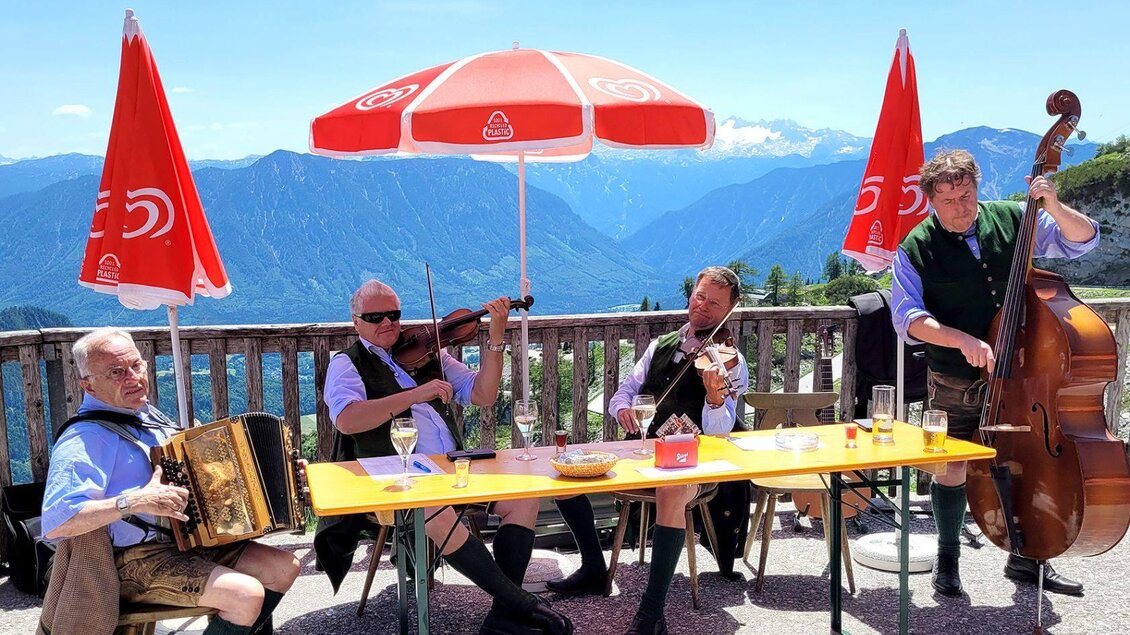 Eine Gruppe von Musikern spielt traditionelle Instrumente unter einem Sonnenschirm mit Blick auf die Berge. Die Stimmung ist fröhlich und einladend. | © RHuber
