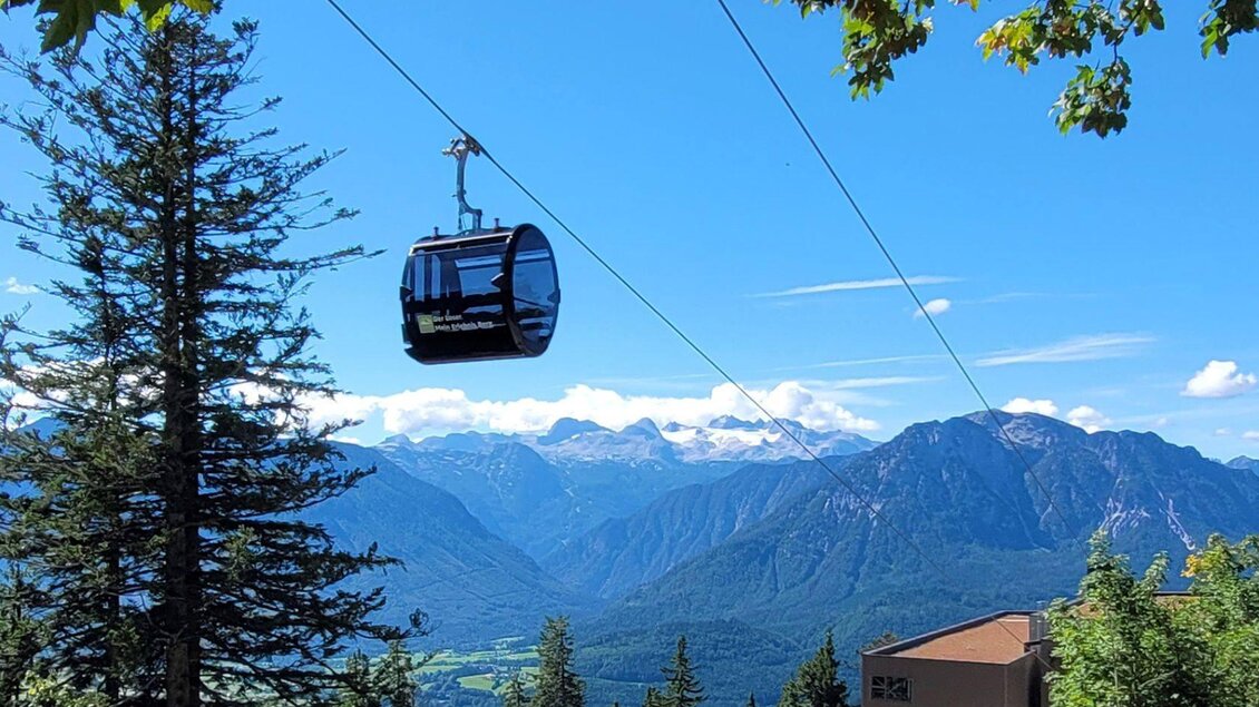 Eine Seilbahn schwebt über ein malerisches Bergpanorama mit grünen Wäldern und schneebedeckten Gipfeln im Hintergrund. Der Himmel ist klar und blau. | © Rudolf Gasperl