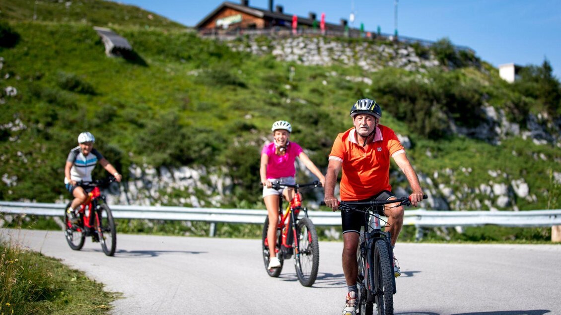 Drei Radfahrer fahren einen bergigen Weg entlang. Im Hintergrund sind grüne Wiesen und eine Almhütte zu sehen. | © TVB Ausseerland - Salzkammergut-Tom Lamm