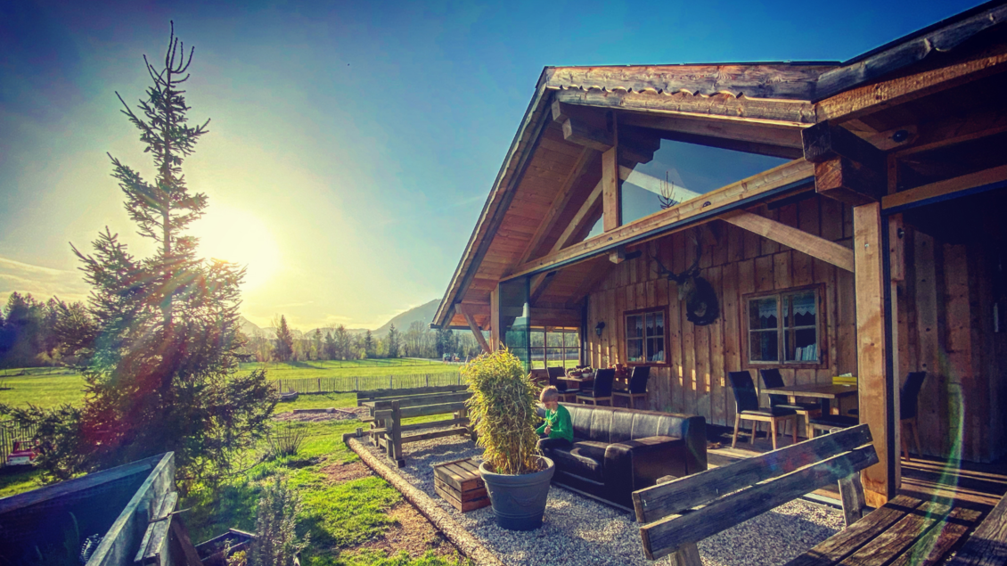 Ein gemütliches Holzhaus mit einer Terrasse in der Abendsonne. Im Vordergrund sind Pflanzen und eine Sitzgelegenheit zu sehen. | © Steirerhütte