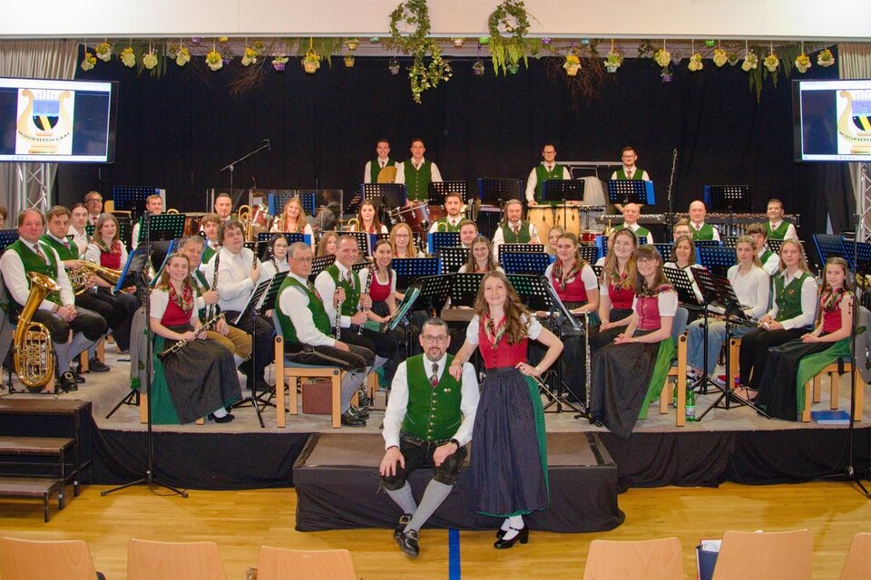 Eine große Blaskapelle sitzt auf einer Bühne in traditioneller Tracht. Im Hintergrund sind Bildschirme mit dem Logo der Kapelle zu sehen. | © Manfred Rohrer