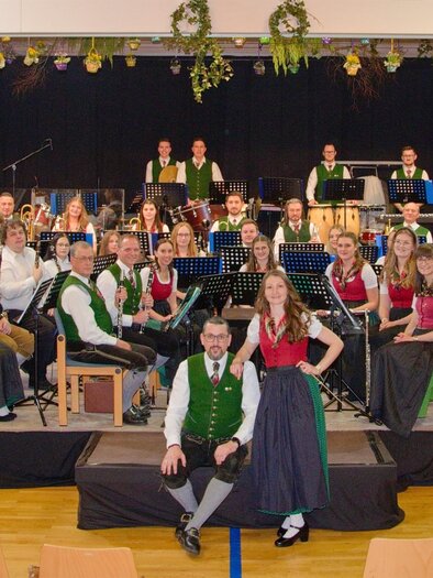 A large brass band is seated on a stage in traditional attire. Screens with the band's logo are visible in the background. | © Manfred Rohrer