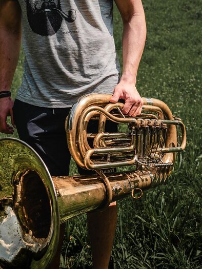 A person is holding a shiny tuba in a green field. In the background, tall grasses can be seen. | © Archiv TV Gesäuse