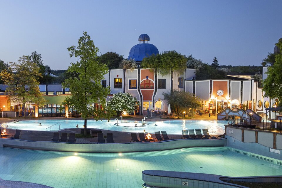 Ein schöner Außenbereich mit einem Pool und einer markanten Architektur. Der Himmel ist klar, und die Umgebung ist von Bäumen umgeben. | © Rogner Bad Blumau © Hundertwasser Architekturprojekt