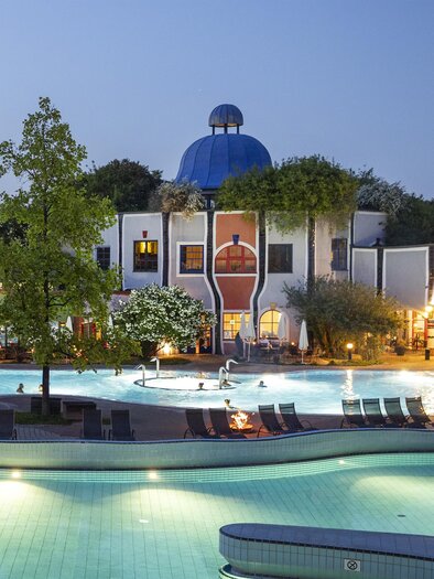 Ein schöner Außenbereich mit einem Pool und einer markanten Architektur. Der Himmel ist klar, und die Umgebung ist von Bäumen umgeben. | © Rogner Bad Blumau © Hundertwasser Architekturprojekt