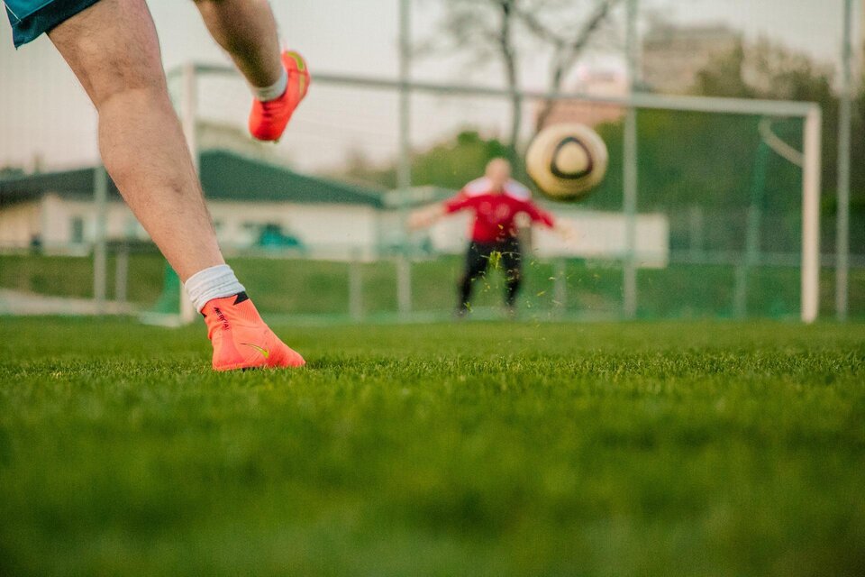Ein Spieler tritt einen Fußball auf einem grünen Spielfeld. Im Hintergrund steht ein Torwart in roter Ausrüstung. | © Pixabay