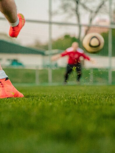 Ein Spieler tritt einen Fußball auf einem grünen Spielfeld. Im Hintergrund steht ein Torwart in roter Ausrüstung. | © Pixabay