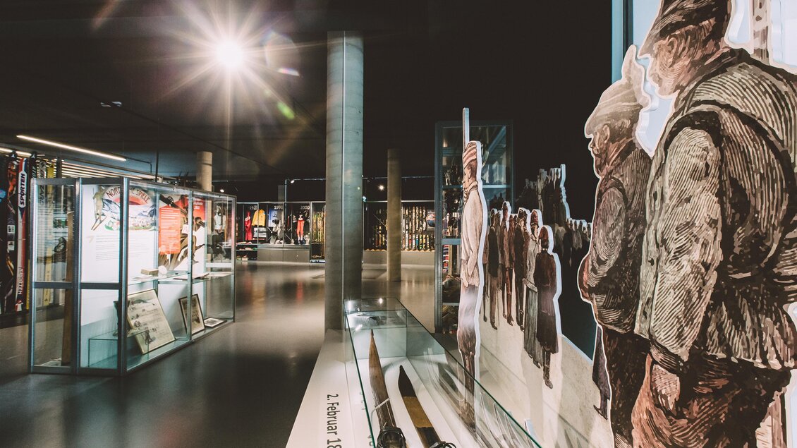 Ein modernes Museum mit Ausstellungsstücken in Vitrinen und Wandmalereien von Menschen. Die Beleuchtung schafft eine einladende Atmosphäre. | © nicoleseiser.at