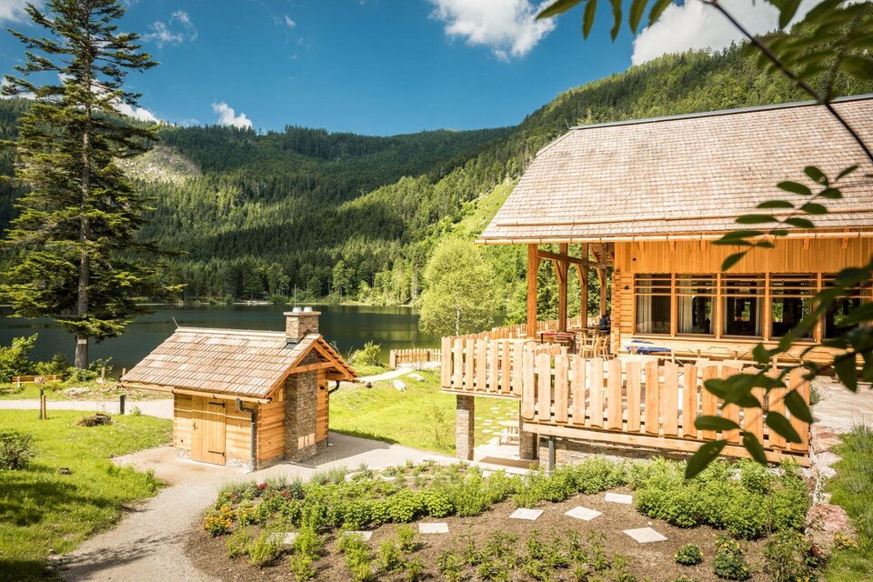 Ein idyllisches Holzhaus mit einer großen Terrasse, umgeben von grüner Landschaft und einem klaren See. Im Hintergrund erheben sich bewaldete Berge unter einem blauen Himmel. | © Genuss am See_ Dieter Sajovic