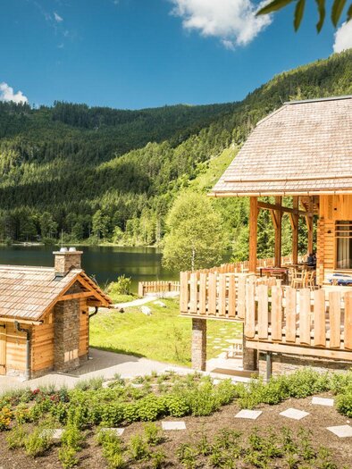 An idyllic wooden house with a large terrace, surrounded by green countryside and a clear lake. In the background, wooded mountains rise under a blue sky. | © Genuss am See_ Dieter Sajovic