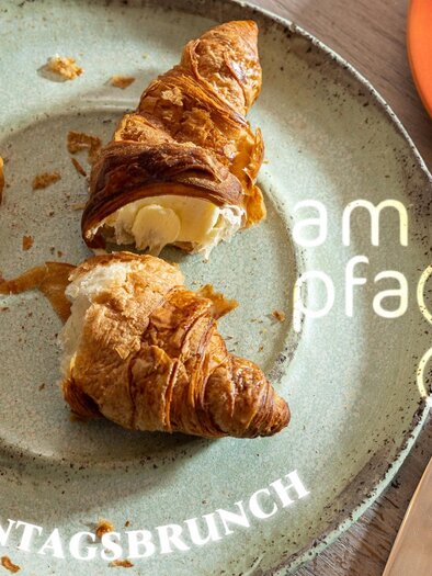 Ein frischer Croissant auf einem Teller, teilweise mit Butter gefüllt. Der Teller ist rustikal und der Hintergrund ist gemütlich. | © kirchgasser_photography