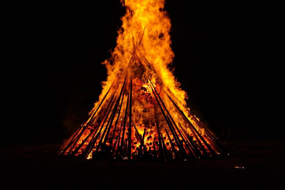 Ein großes Lagerfeuer brennt hell in der Dunkelheit. Die Flammen lodern hoch zwischen den hölzernen Stangen.