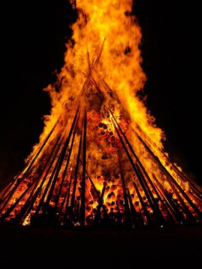 Ein großes Lagerfeuer brennt hell in der Dunkelheit. Die Flammen lodern hoch zwischen den hölzernen Stangen.