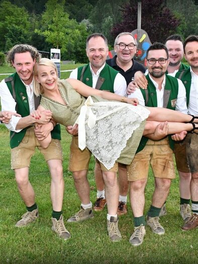 Eine fröhliche Gruppe von Männern in traditioneller Tracht steht zusammen mit einer Frau in einem Kleid. Alle lächeln, während die Frau von einigen Männern gehalten wird. | © DALUIS