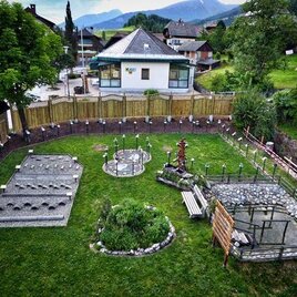 Ein gepflegter Garten mit verschiedenen Steinfeldern und einem kleinen Gewässer. Im Hintergrund steht ein Gebäude und die Umgebung ist von Bäumen und Bergen umgeben.