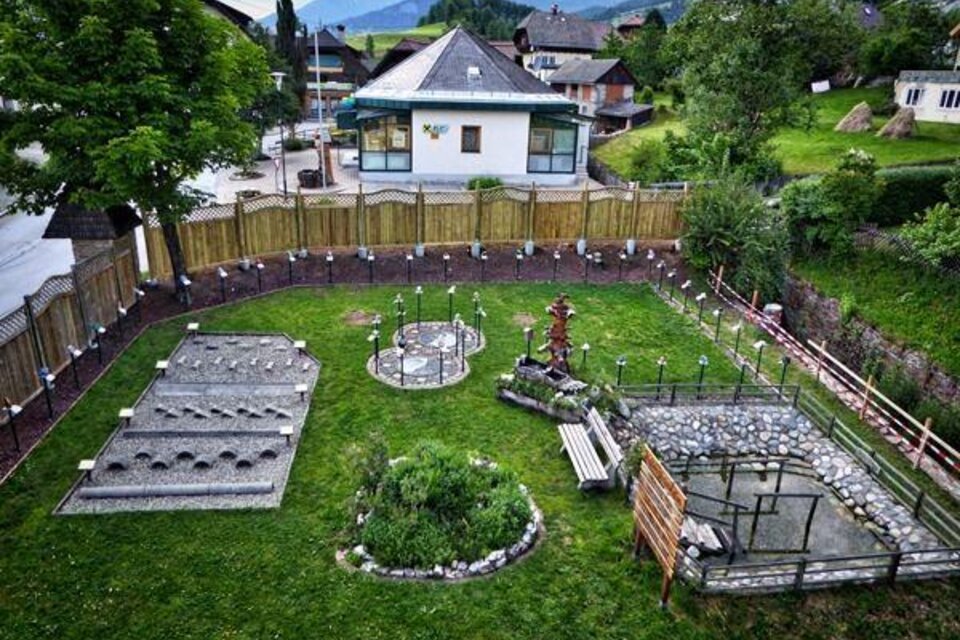 Ein gepflegter Garten mit verschiedenen Steinfeldern und einem kleinen Gewässer. Im Hintergrund steht ein Gebäude und die Umgebung ist von Bäumen und Bergen umgeben.