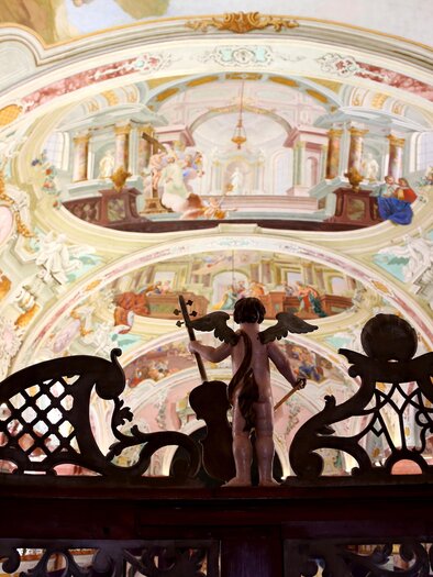Eine kunstvoll verzierte Decke mit barocken Fresken. Im Vordergrund stehen zwei Engelstatuen, die den Blick auf die Malerei lenken. | © Stift Rein