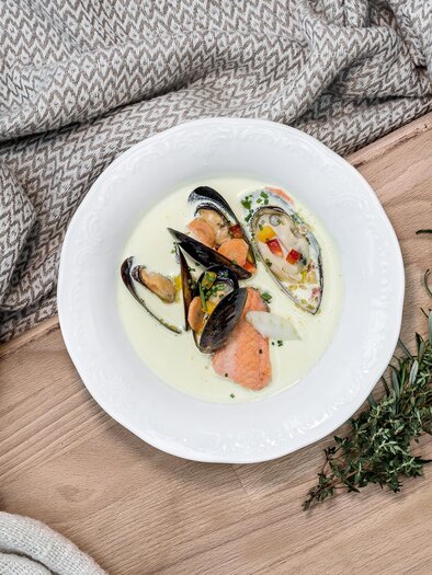 Ein schönes Gericht mit Muscheln und Lachs in einer cremigen Sauce. Das Essen ist auf einem weißen Teller angerichtet und von einem gemütlichen Tuch umgeben. | © Fischrestaurant Kulmer