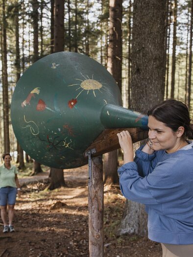 Eine Frau schaut durch ein großes, grün bemaltes Hörrohr im Wald. Im Hintergrund steht eine andere Person und genießt die Natur. | © nicoleseiser.at