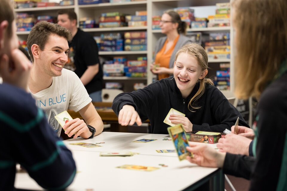 Eine Gruppe von Jugendlichen spielt lächelnd an einem Tisch mit Gesellschaftsspielen. Im Hintergrund sind viele Spiele im Regal zu sehen. | © Spielefreunde