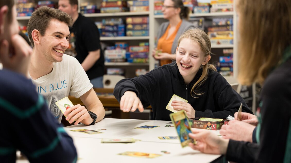 Eine Gruppe von Jugendlichen spielt lächelnd an einem Tisch mit Gesellschaftsspielen. Im Hintergrund sind viele Spiele im Regal zu sehen. | © Spielefreunde