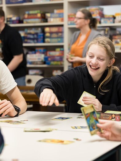 Eine Gruppe von Jugendlichen spielt lächelnd an einem Tisch mit Gesellschaftsspielen. Im Hintergrund sind viele Spiele im Regal zu sehen. | © Spielefreunde