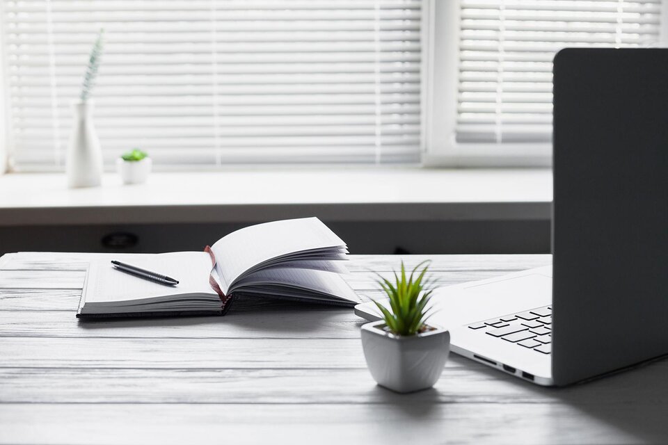 Ein Schreibtisch mit einem geöffneten Notizbuch, einem Stift und einem Laptop. Im Hintergrund sind kleine Pflanzen und ein Fenster mit Jalousien zu sehen. | © Bild: freepik