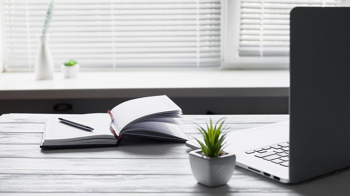 Ein Schreibtisch mit einem geöffneten Notizbuch, einem Stift und einem Laptop. Im Hintergrund sind kleine Pflanzen und ein Fenster mit Jalousien zu sehen. | © Bild: freepik