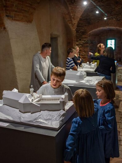 Egy múzeum látogatókkal, akik történelmi épületek makettjeit nézik. Néhány gyermek egy asztalnál áll, és érdeklődve nézik a makettet. | © Gartenschloss Herberstein