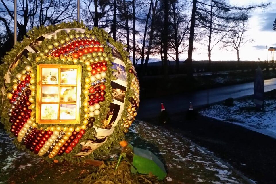 Eine große, runde Weihnachtsdekoration aus bunten Weihnachtskugeln und Äpfeln steht in der Dämmerung. Im Hintergrund sind Bäume und ein Weg sichtbar.
