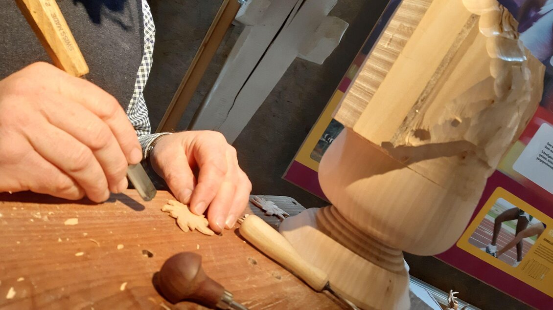 Ein Handwerker schnitzt Holz und hält ein Werkzeug in der Hand. Im Hintergrund sind Informationen über Holzverarbeitung sichtbar.