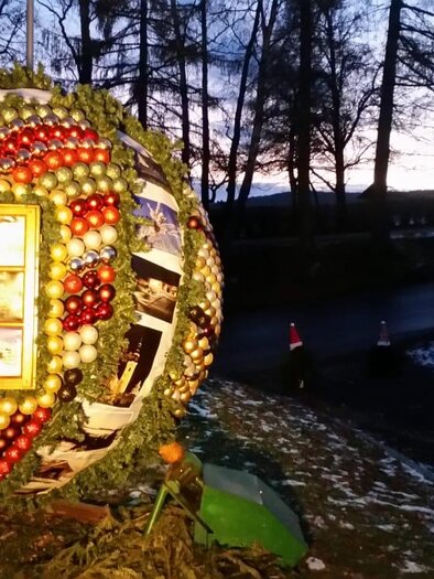 Eine große, runde Weihnachtsdekoration aus bunten Weihnachtskugeln und Äpfeln steht in der Dämmerung. Im Hintergrund sind Bäume und ein Weg sichtbar.