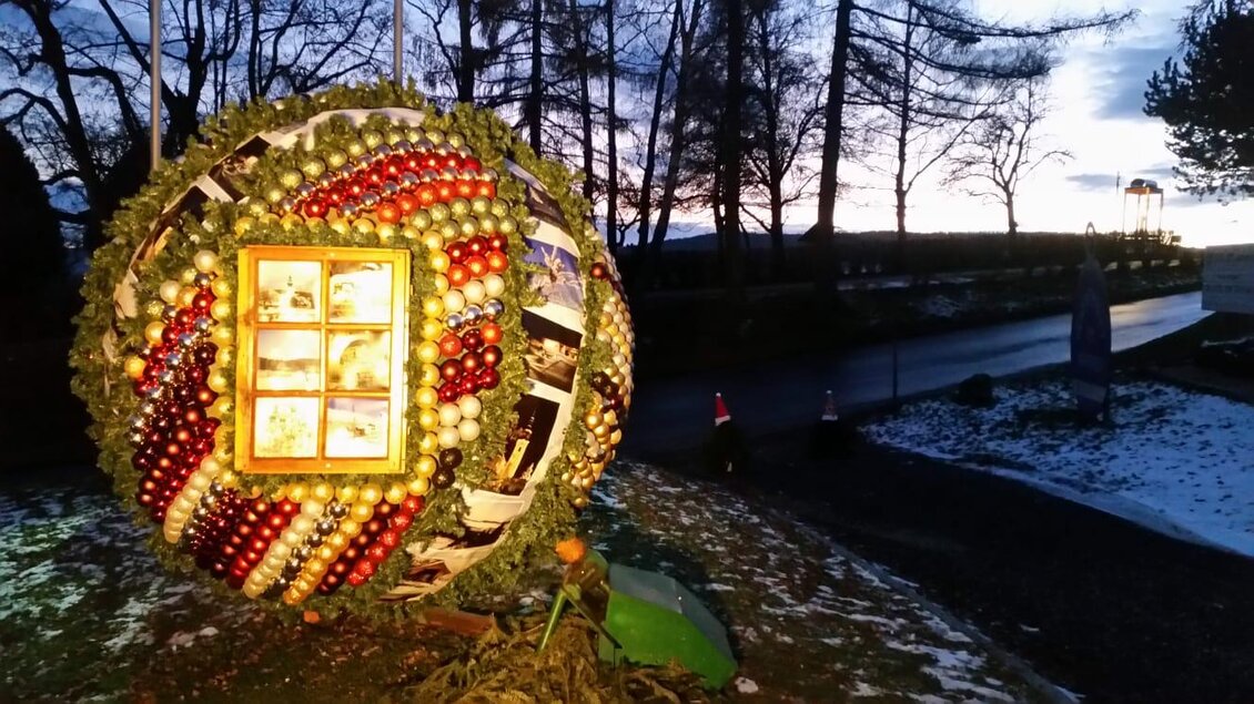 Eine große, runde Weihnachtsdekoration aus bunten Weihnachtskugeln und Äpfeln steht in der Dämmerung. Im Hintergrund sind Bäume und ein Weg sichtbar.
