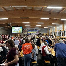 Eine große Menschenmenge feiert in einer Halle mit langen Tischen. Viele tragen traditionsreiche Kleidung und es gibt eine festliche Stimmung. | © USV St. Lorenzen am Wechsel