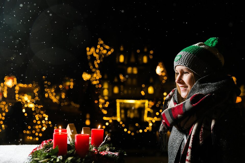 Eine Frau mit einer Mütze steht in einer festlichen Umgebung. Im Hintergrund leuchten Weihnachtslichter und es schneit leicht. | © Lupi Spuma