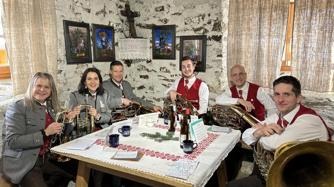 Sechsergruppe von Musikern in traditioneller Tracht sitzt an einem Tisch. Sie halten Instrumente und umgeben sich mit Getränken in einem rustikalen Raum.