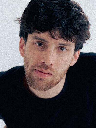 A young man with curly hair is wearing a black T-shirt. He is looking directly at the camera and has a thoughtful expression.