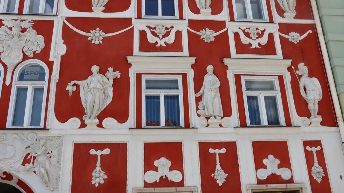 Eine historische Fassade mit rotem Putz und dekorativen weißen Reliefs. Vier Figuren sind prominent abgebildet, umrahmt von Fenstern. | © TVB Erzberg-Leoben