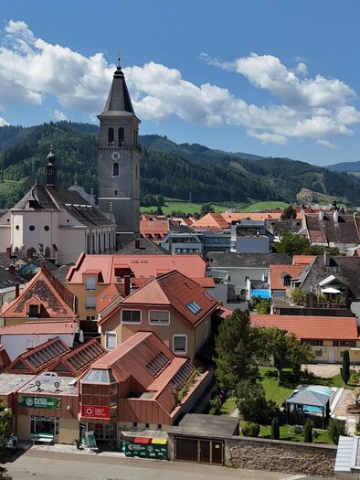 Eine lächelnde Frau steht an einem Balkon mit schöner Aussicht auf eine grüne Landschaft. Sie trägt ein buntes Oberteil und schaut freundlich in die Kamera. | © Erlebnisregion Murtal