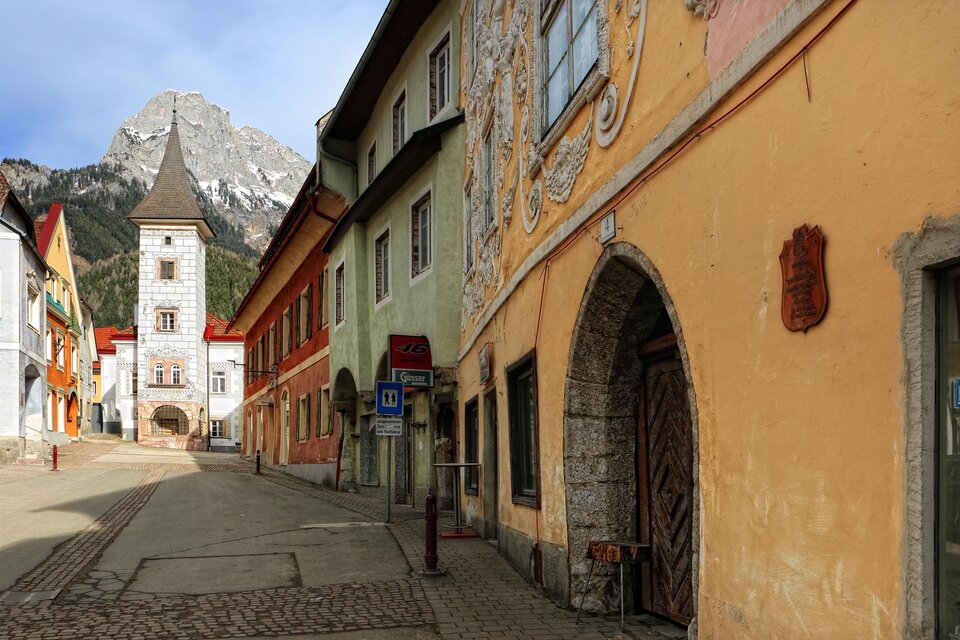 Ein historisches Gebäude mit dekorativen Fassadenmalereien und Fenstern. Im Hintergrund sind Berge zu sehen, bei klarem Himmel. | © Pezmann